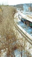 A freight train with containers moves along the rails in the direction of the city in a winter day. Shipping of goods. Aerial view. Vertical video