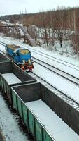 A freight train is passing by rail along rows of containers outside the city in the station area in the winter. Aerial view. Vertical video