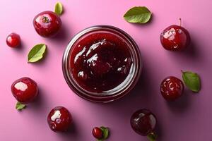Fresh Homemade Cherry Jam in a Glass Jar with Whole Cherries and Leaves on a Pink Background photo