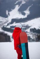 A man in a red jacket with a snowboard stands on a mountainside. photo