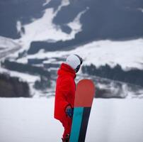 A man in a red jacket with a snowboard stands on a mountainside. photo