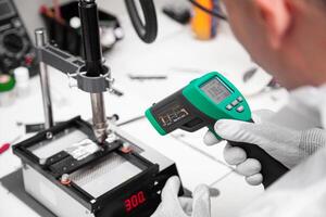 The master measures the temperature of the microchip at the soldering station with a laser thermometer. Service center. Repair of microelectronics. photo
