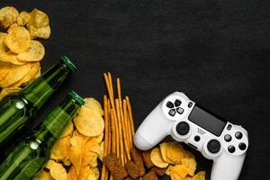 Modern white gamepad among chips, crackers and bottles of beer on a black. Game concept. photo