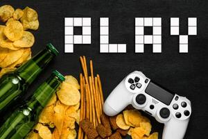 Modern white gamepad among chips, crackers and bottles of beer on a black. Game concept. photo
