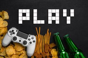 Modern white gamepad among chips, crackers and bottles of beer on a black. Game concept. photo