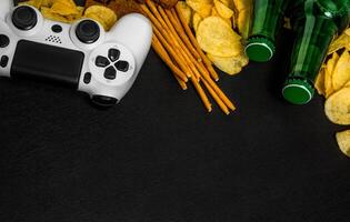 Modern white gamepad among chips, crackers and bottles of beer on a black. Game concept. photo