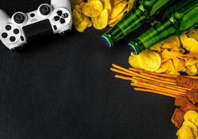 Modern white gamepad among chips, crackers and bottles of beer on a black. Game concept. photo