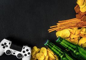 Modern white gamepad among chips, crackers and bottles of beer on a black. Game concept. photo