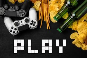 Modern white gamepad among chips, crackers and bottles of beer on a black. Game concept. photo