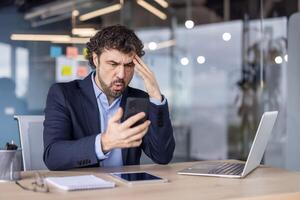 Businessman in suit experiencing frustration while looking at phone in modern office. Emotional reaction to message or email, working with laptop and tablet at desk. photo