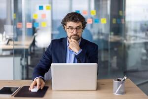 Businessman in suit working on laptop at modern office. Professional focused on tasks with sticky notes in background indicating organization and planning. Ideal for business corporate photo