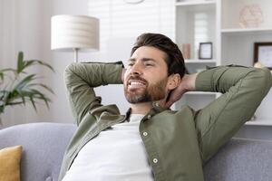 Relaxed man sitting on sofa with hands behind head, enjoying leisure time at home. Man smiling while resting and feeling stress-free indoors. photo