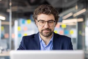Business professional with glasses and beard working on laptop in modern office space, focused and concentrated photo