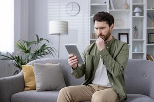 Man seated on sofa reading tablet at home. Thoughtful expression while using digital device. Modern living room background. Concept of technology, leisure, home office, remote work, reading, phone photo