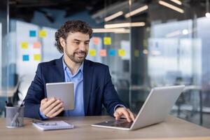 Confident businessman working with digital tablet and laptop in modern office. Professional wearing formal attire, focusing on work tasks, surrounded by sticky notes on glass walls photo