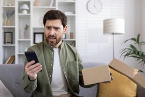 Man looking at phone with shocked expression while holding empty package box. Concept of online shopping disappointment, unexpected delivery issues, or receiving wrong items photo