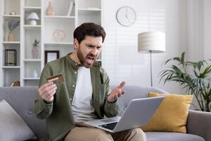 Man expressing frustration with online payment holding credit card and using laptop on couch at home. Concept of online shopping issues, payment failure, and internet banking problems. photo