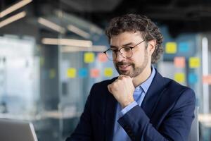 Confident businessman working on computer in modern office, focused on project with colorful sticky notes in background. Professional and reflective mindset, dressed in suit and glasses. photo
