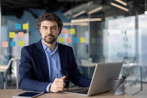 Confident businessman working on laptop in modern office with glass walls and post-it notes. Professional setting for corporate planning, strategy, or execution. photo