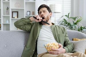 A bored young man yawning while holding a TV remote and a bowl of popcorn, sitting on a sofa in a living room setting. photo