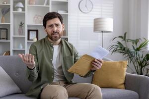 Man sitting on sofa holding envelope looking worried signifying bad news. Home interior setting with modern furniture. Image conveys concern, uncertainty, and financial stress. photo