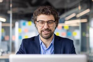 Smiling businessman working on laptop in modern office setting with glass walls and sticky notes. Professional wearing glasses focusing on computer screen photo
