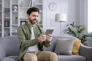 Smiling man using tablet while sitting on sofa at home. Happy individual enjoying modern technology, engaging in leisure time or remote work. Light and cozy home interior. Android and iOS compatible. photo