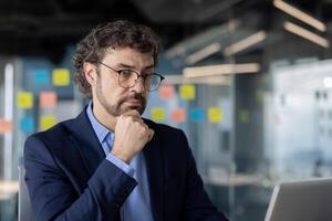 Businessman wearing glasses deep in thought while working in modern office. Professional setting with focus on contemplation and strategy. photo