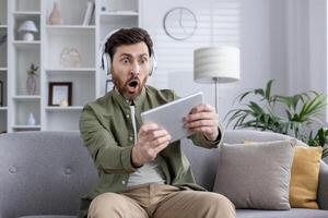 Man sitting on couch wearing headphones, using tablet for game. Expresses excitement and surprise, likely from gaming experience. Reflects themes of technology, entertainment, modern lifestyle. photo