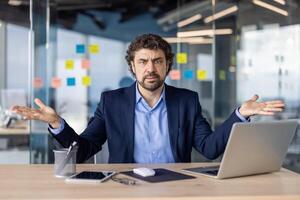 Confused businessman sitting at desk in modern office, shrugging with both hands. Man in suit with laptop, tablet, mouse, and stationery, expressing uncertainty or confusion. photo