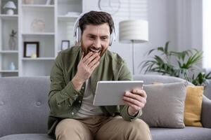 Man sitting on sofa wearing headphones while watching a funny on tablet. He laughs and touches his face, engrossed in content. Relaxed home setting with modern decor. photo