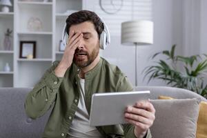 Man wearing headphones using tablet with frustrated expression sitting on couch at home. Feeling of stress, tiredness, and technology issues. photo