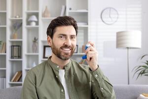 Man using inhaler for asthma relief sitting at home smiling. Person demonstrating proper use of medical device for respiratory condition. Concept of healthcare, respiratory therapy, chronic disease. photo