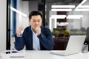 Young Asian male businessman sitting at desk in office and suffering from severe cough and asthma attack, holding napkin and covering mouth with hand. photo