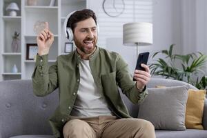 Man wearing headphones holding phone, smiling and pointing while sitting on couch. Enjoying call, music, or online interaction indoors. Casual, relaxed atmosphere capturing positive emotions. photo