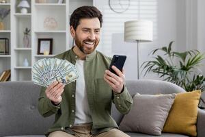 Smiling man sitting on a couch, displaying a spread of dollar bills while happily using his mobile phone at home. photo