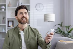 Satisfied man holding air conditioner remote control in hands, smiling indoors. Man adjusting temperature with remote. Concept of comfort, technology, and wellbeing. photo