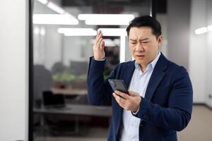 Man in business suit inside office with phone in hands, mature businessman received online notification message with bad news, boss uses app on smartphone, upset received rejection error. photo