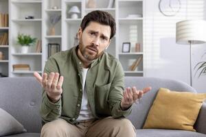 Man sitting on sofa expressing confusion during call using phone in living room. Man making hand gestures looking at camera, feeling uncertain and perplexed. photo