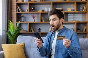 Man sitting on a couch looking shocked while holding a smartphone and a credit card, possibly after seeing unexpected charges. photo