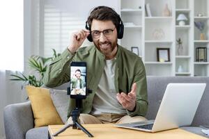 Smiling man with headphones having an online call using his laptop and smartphone while sitting on the sofa at home. photo