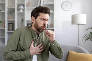 Man with beard, sitting on couch, experiencing chest pain and coughing. Potential health issues like heart problems or respiratory conditions shown. Casual home setting, daytime environment. photo