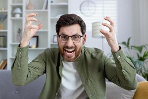 Man in casual outfit with glasses showing frustration and anger in a home setting. Emotions of stress and anxiety clearly visible. photo