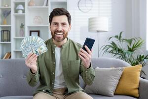 Portrait of happy young man sitting on sofa at home and enjoying financial success on camera, holding mobile phone and cash money. photo