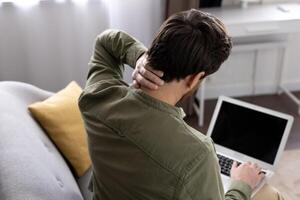 Man experiencing neck pain while using laptop at home. Discomfort from prolonged sitting and computer use. Concept of workplace ergonomics, health, and stress from technology use. photo