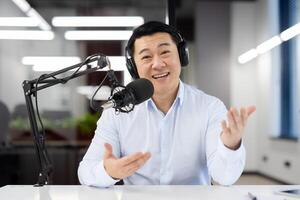 Young Asian man working in office, sitting at desk wearing headphones and in front of desk with microphone and talking to camera via call. conducts an online meeting, records business training. photo