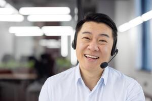 Webcam view, Asian support worker in shirt with headset smiling and looking at camera, businessman working inside office, consulting clients remotely, remote meeting with colleagues partners. photo