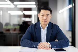 Portrait of a confident Asian businessman seated at a table in a well-lit, contemporary office environment, looking at the camera with focus. photo