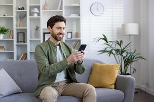 Satisfied mature man sitting on sofa in living room, using app on phone, freelancer holding smartphone, reading and typing text message, browsing social media. photo