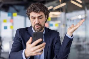 Confused businessman in suit looking at cell phone in modern office setting. Man appears perplexed or frustrated while holding smartphone, likely dealing with unexpected news or information. photo